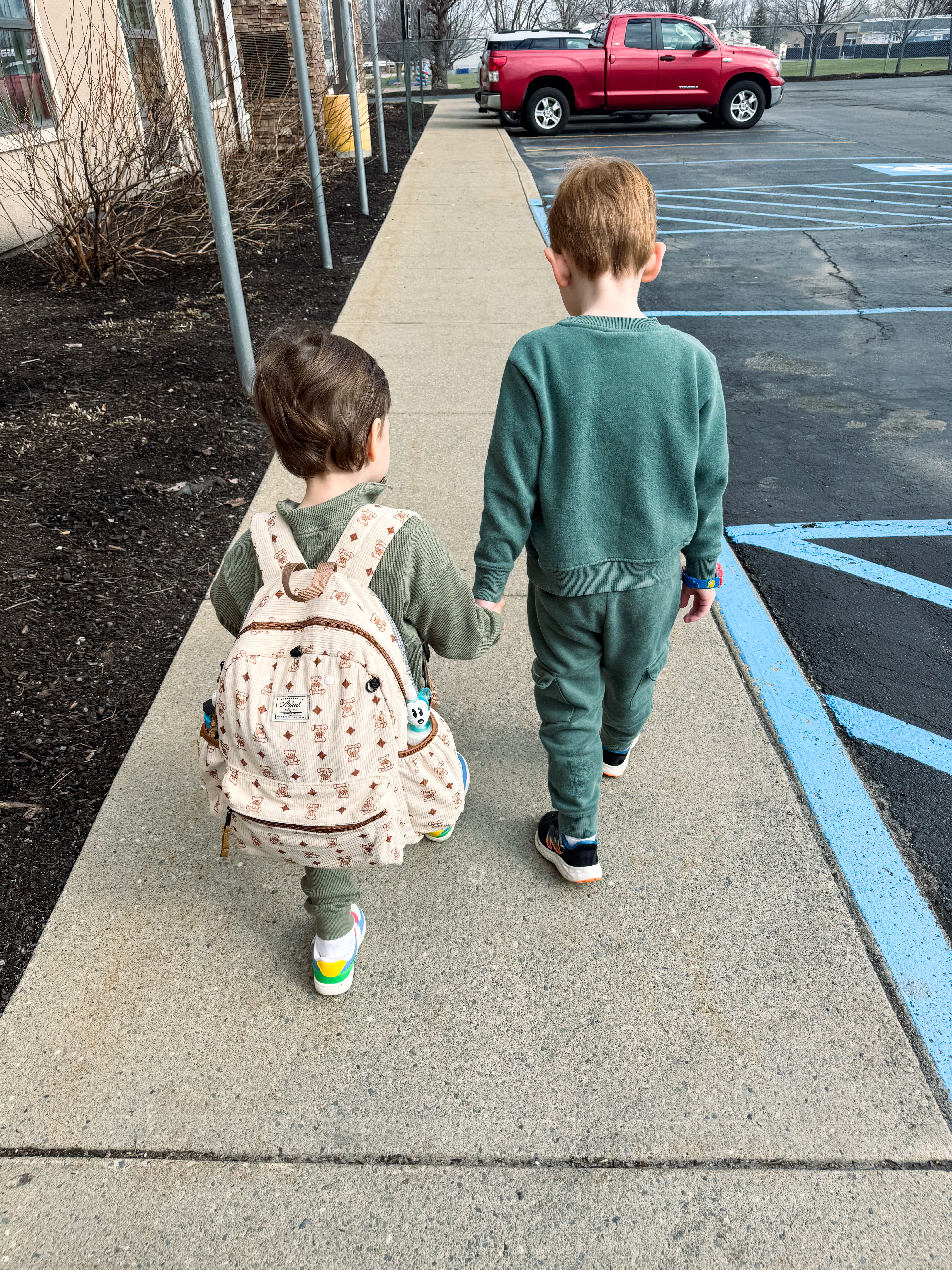 Two children walking hand in hand on a sidewalk, one with a backpack and the other in matching attire.